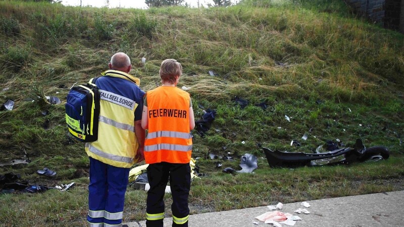 Ein Seelsorger und ein Mitglied der Feuerwehr an einer Unfallstelle auf der Autobahn 9 vor einer Böschung, die mit Trümmerteilen übersäht ist.