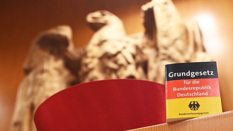 Vor dem Bundesadler im Sitzungssaal des Bundesverfassungsgericht steht eine Miniaturausgabe des Grundgesetz sowie ein Barett eines Bundesverfassungsrichters.